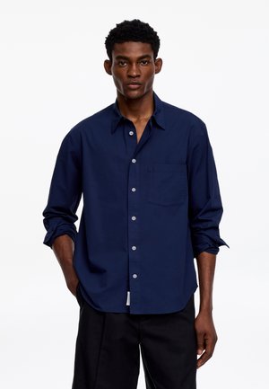 Homme aux cheveux courts et bouclés portant une chemise bleu marine à boutons avec manches retroussées et un pantalon noir, debout les mains dans les poches.