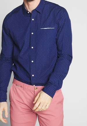 Homme portant une chemise à manches longues bleu marine à motifs et un pantalon rose saumon, debout devant un fond uni.