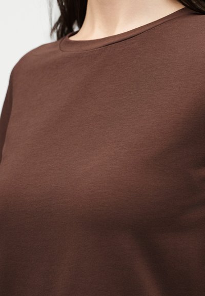 Κοντινό πλάνο ενός ατόμου που φοράει απλό καφέ t-shirt με στρογγυλή λαιμόκοψη, με ίσια σκούρα μαλλιά που φαίνονται πάνω από τη λαιμόκοψη.