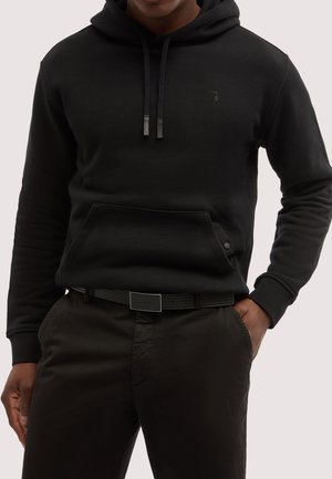 Uomo con felpa nera con tasca frontale, coulisse e piccolo logo ricamato, abbinata a pantaloni neri e cintura, mano in tasca.