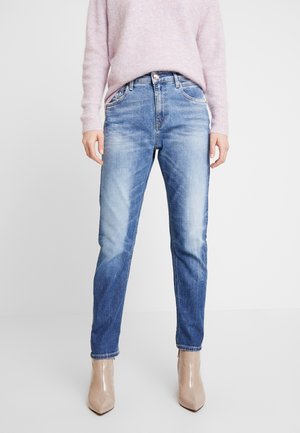 Žena oblečená v světle růžovém svetru, modrých vybledlých džínech s rovnými nohavicemi a béžových členkových botách se špičatou špičkou stojící na bílém pozadí.