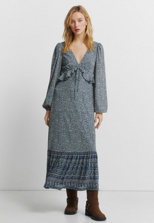 Długa, granatowa sukienka z białym wzorem paisley, z głębokim dekoltem w serek, marszczonym pasem i falbanami na dole. Do tego brązowe botki za kostkę.