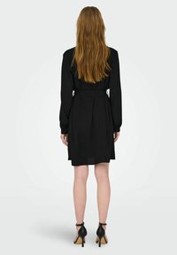 Black dress with long sleeves, belted waist, pleated back, knee-length, paired with black heeled sandals. Smooth texture.