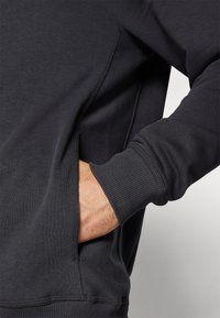 Mørkegrå sweatshirt med ribbede manchetter, sidelomme og glat tekstur, der viser et nærbillede af en hånd, der hviler i lommen.