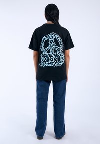 Camiseta de algodón negra con un gráfico de signo de paz azul claro y texto en la parte posterior. Corte holgado con mangas cortas, combinada con pantalones de mezclilla azules.
