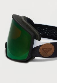 Slēpošanas brilles ar zaļu atstarojošu lēcu, melnu putu polsterējumu un regulējamu melnbaltu jostu, uz kuras ir brūna ādas logotipa birka.