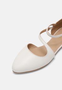 Chaussure plate blanche à bout pointu avec une semelle intérieure beige et une fine bride traversant le pied, sur un fond blanc uni.