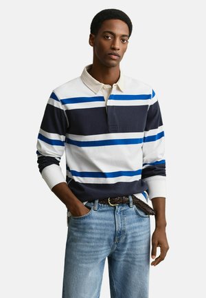 Jonge man die een rugbyshirt met lange mouwen draagt met blauwe, marineblauwe en witte horizontale strepen, gecombineerd met lichtblauwe spijkerbroek en een gevlochten bruine riem.