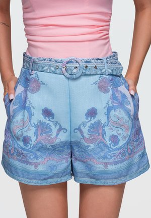 Femme portant un short bleu clair à motif floral avec une ceinture assortie et un haut rose sans manches, mains visibles dans les poches.
