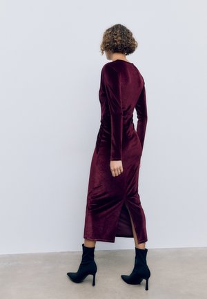 Burgundy samettimekko pitkillä hihoilla, rypytetty sivudettaali ja korkea halkio, yhdistetty mustiin korkokenkiin.