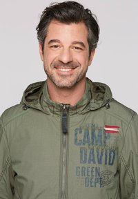 Green zip-up jacket with a hood, featuring text and decorative stitching on the front. Material appears lightweight with a slightly textured finish.