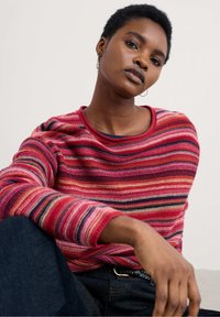 Pulover rayat în nuanțe de roșu, roz și negru, cu guler rotund, țesătură texturată și croială lejeră, asortat cu blugi întunecați.