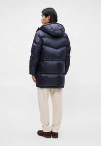 Casaco puffer azul-marinho com capuz, apresentando um design acolchoado e punhos elásticos. Usado sobre calças bege e sapatos castanhos.