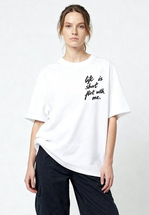 Jonge vrouw met het haar in een staart, gekleed in een wit oversized T-shirt met zwarte tekst "Life is short flirt with me." en een zwarte broek.