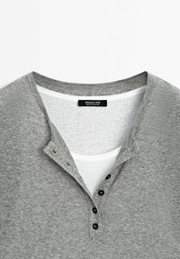 Maglietta henley grigia con texture a costine, tre bottoni neri al colletto e uno strato interno bianco visibile all'escollatura. Etichetta del marchio inclusa.