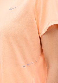 Κοντινό πλάνο ενός ροδακινί κοντομάνικου πουκάμισου με μικρό γκρι λογότυπο Nike Swoosh και γκρι οπές εξαερισμού κοντά στη ραφή του πλάγιου μέρους.