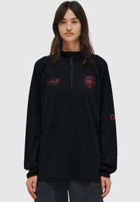 Svart fleece-pullover med dragkedja i halsen, har röda broderade logotyper på bröstet och ärmen samt en strukturerad textil. Långa ärmar med manschetter.