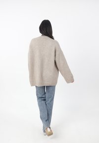 Jersey de punto beige de gran tamaño con cuello alto, ajuste relajado y puños acanalados, combinado con jeans rectos de tiro claro y sandalias de tacón transparente.
