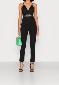 Kvinna i en svart halterneck jumpsuit med spetsdetaljer, svarta klackar med ankelsrem och hållande en liten grön handväska med en guldkedja.