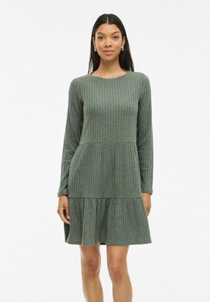 Femme aux longs cheveux noirs portant une robe tricotée côtelée verte à manches longues avec une jupe à volants, se tenant devant un fond blanc uni.