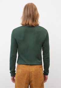 Camicia verde scuro a maniche lunghe con una trama liscia, abbinata a pantaloni in velluto a coste beige con tasche posteriori e cuciture discrete.