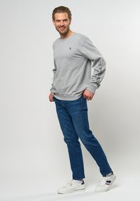 Grå hettegenser med rund hals og lange ermer, med logo på brystet og tekst på ermet. Kombinert med blå jeans og hvite joggesko.