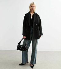 Cappotto doppiopetto nero, jeans a gamba larga sbiaditi, décolleté nere con punta, e una borsa nera con dettagli a borchie. Tessuto morbido e design strutturato.
