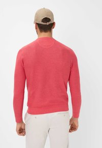 Roter Pullover mit langen Ärmeln und Rundhalsausschnitt, aus leichtem Strickstoff. Hellbeige Mütze, rückwärts getragen.