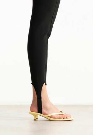 Onderbeen met zwarte beenstukken met beugel gecombineerd met bleekgele kitten heel sandalen op een lichte vloer en witte achtergrond.