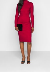 Red ribbed midi dress with a turtleneck and long sleeves, paired with a black clutch and open-toe black heeled mules. Features a fitted silhouette.