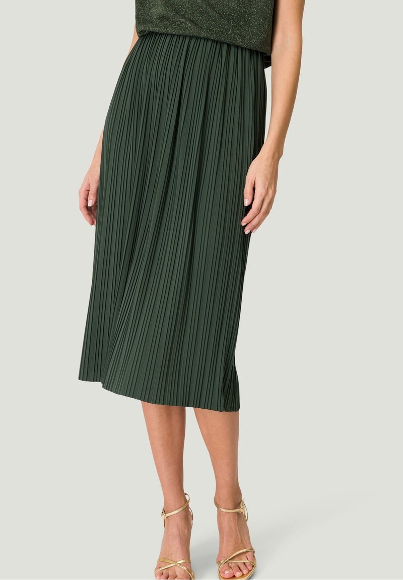 Femme portant une jupe midi plissée vert foncé et des sandales à talons hauts dorées à brides, debout devant un fond clair uni.