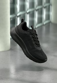 Baskets Calvin Klein noires à lacets, flottant au-dessus d'un sol gris poli près d'un mur en blocs de verre et d'un pilier blanc.