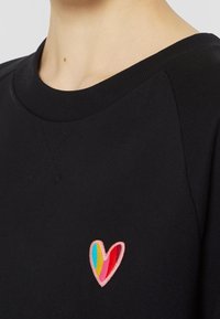 Gros plan sur une personne portant un sweat-shirt noir avec un petit patch brodé en forme de cœur en rose, rouge, bleu et jaune sur la poitrine.
