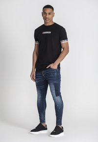 Camiseta negra de manga corta con acentos de cuadros en las mangas, combinada con jeans de denim oscuro de corte ajustado, con un acabado desgastado y detalles salpicados de blanco.