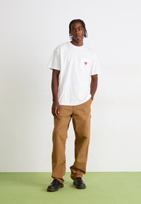 Young man standing with hands in pockets, wearing white t-shirt with red heart on pocket, brown pants, and black shoes on green floor.