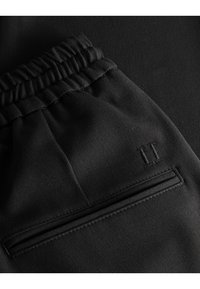Schwarze Jogginghosen aus strukturiertem Stoff, mit einem elastischen Bund und einem Reißverschluss-Taschedetail. Genähter Akzent mit subtiler Logo-Darstellung.