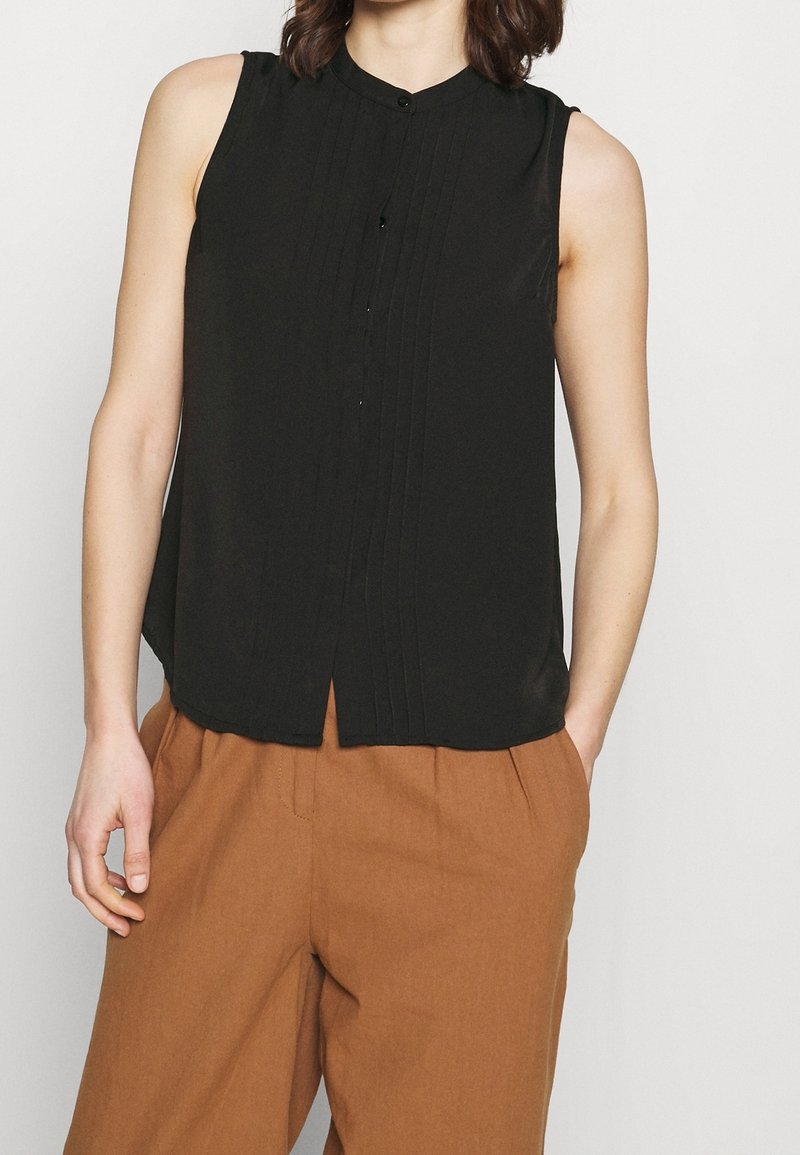 Blouse noire sans manches avec plis et boutons sur le devant, portée avec un pantalon large marron taille haute, mains détendues sur les côtés.