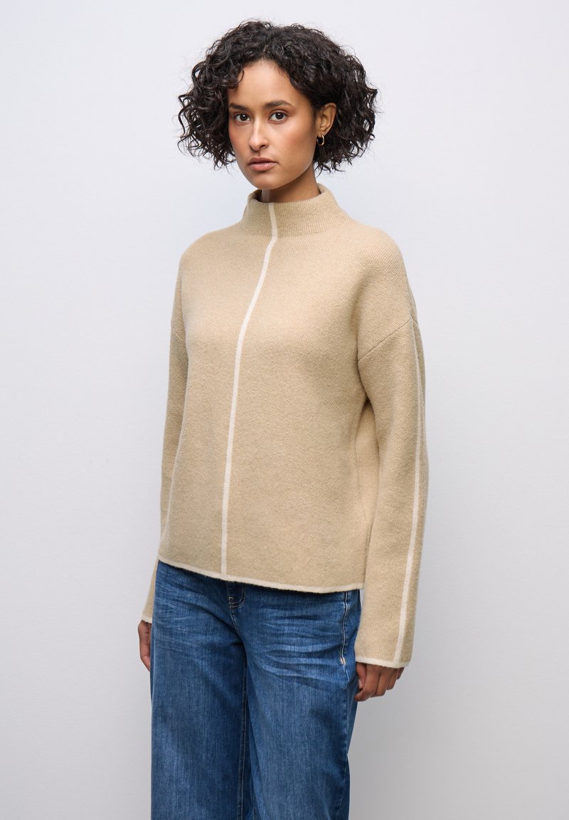 Beige Pullover mit hohem Kragen aus strukturiertem Strick, versehen mit einem vertikalen cremefarbenen Streifen, entspannter Passform und überschnittenen Schultern, kombiniert mit blauen Jeans.