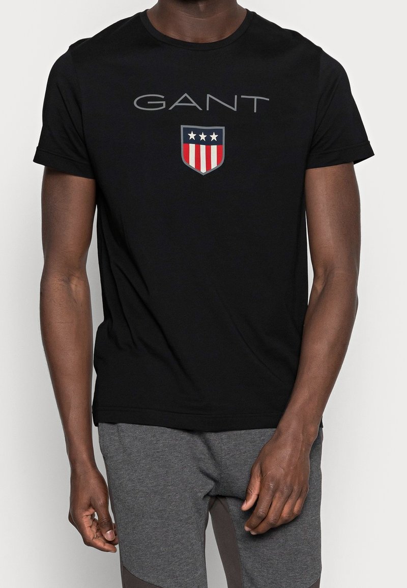 Camiseta negra de manga corta con el texto "GANT" y un logo de escudo con tres estrellas y franjas rojas, llevada por una persona con pantalones grises.