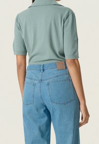 Vihreä neulottu lyhythihainen poolopaita, yhdistetty vaaleansinisiin korkeavyötäröisiin farkkuihin, joissa on takataskut ja ruskea nahkainen merkkiläppä.