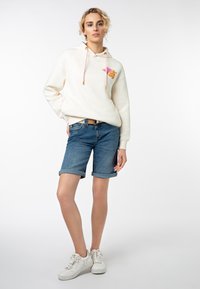 Gekapuerde crèmekleurige sweatshirt met roze trekkoorden en kleurrijke print, gecombineerd met opgerolde blauwe denim shorts en witte sneakers.