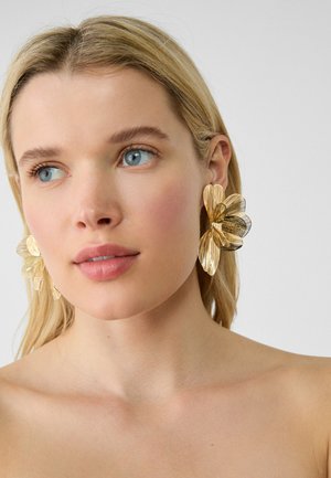 Donna bionda con occhi azzurri che indossa grandi orecchini a forma di fiore metallici dorati su uno sfondo chiaro e semplice.