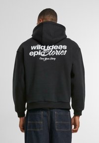 Schwarzer Kapuzenpullover mit Kängurutasche, auf der Rückseite mit großen weißen Buchstaben bedruckt. Der Stoff ist weich und hat gerippte Bündchen und Saum.