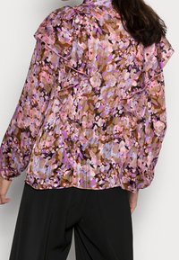 Blusa trasparente con motivo floreale multicolore, con rouches sulle spalle, maniche lunghe e un taglio leggermente morbido. Finita con polsini elasticizzati.