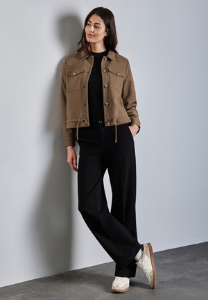 Bruine cropped jas met twee borstzakken, gecombineerd met zwarte broek met wijde pijpen en witte sneakers, tegen een grijze achtergrond.