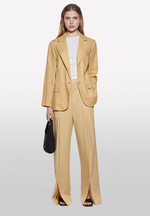 Blazer giallo in lino e pantaloni abbinati con spacchi, abbinati a un top bianco a coste, una borsa intrecciata nera e sandali neri con tacco.