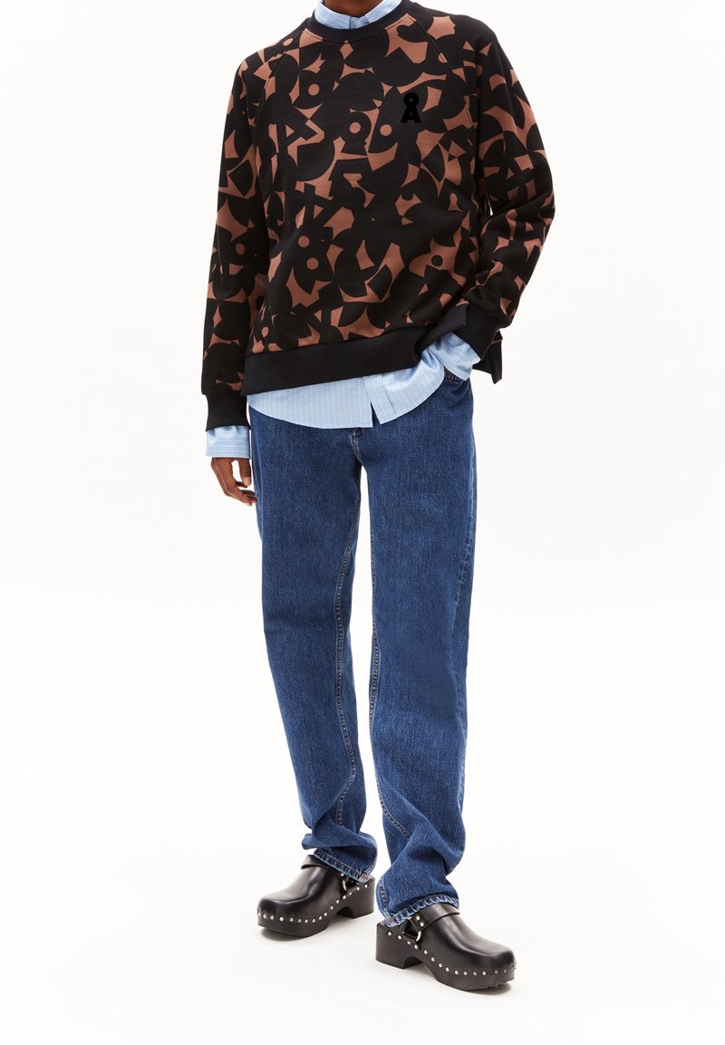 Personne portant un pull à motif abstrait noir et marron sur une chemise bleu clair, un jean bleu et des sabots noirs cloutés à talons.