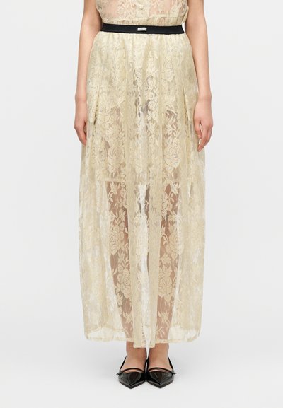 Μακριά φούστα από δαντέλα σε απαλή κίτρινη απόχρωση με floral σχέδια, με μαύρη μέση στη μέση. Διαφανής υφή με στρωματοποιημένο σχέδιο, φορεμένη με μαύρα μυτερά παπούτσια.
