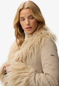 Vêtement d'extérieur en velours côtelé beige avec un col en fausse fourrure texturé, long et de couleur claire. Présente une silhouette ajustée et un logo de marque discret sur la manche.