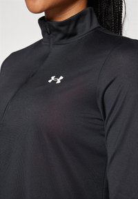 Schwarzes athletisches Half-Zip-Oberteil aus glattem Stoff, mit kleinem weißem Under Armour-Logo auf der linken Brust und einem hochgeschlossenen Kragen.